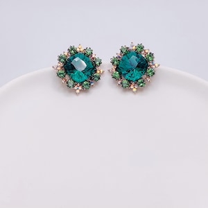 May include: A pair of emerald green stud earrings. Each earring has a large, faceted, round-cut green gemstone surrounded by smaller green and iridescent crystals. The earrings are set on a white surface.