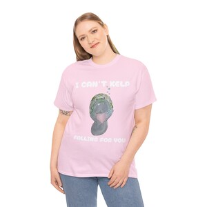 May include: Pale pink t-shirt with the text "I CAN'T KELP FALLING FOR YOU". The shirt features a watercolour illustration of a manatee holding a pink heart. The manatee is grey and green.