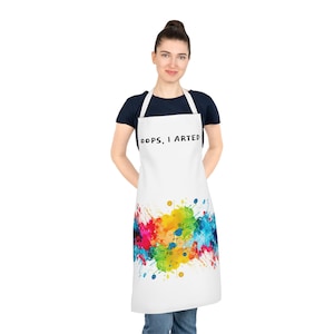 Puede incluir: Delantal blanco con un diseño de salpicaduras de pintura de colores y el texto "Oops, I arted" en negro.
