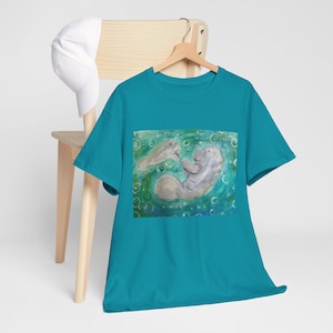 May include: Teal t-shirt with a watercolour painting of two manatees in a watery scene with bubbles. The manatees are grey and white, set against a green and blue background. The shirt is displayed on a wooden hanger.