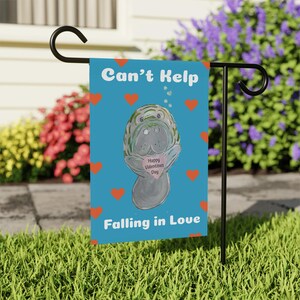 May include: A blue garden flag featuring a cartoon manatee holding a pink heart with the text "Happy Valentines Day". The flag has the text "Can't Help Falling in Love" and orange hearts.