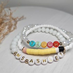 May include: Two white beaded bracelets with the word "TEACH" spelled out in colourful beads on one bracelet and the name "ASHLEY" spelled out in white beads on the other bracelet.