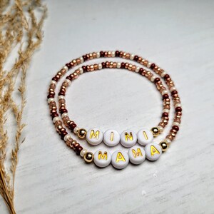 May include: Two beaded bracelets. The top bracelet spells out "MINI" and the bottom bracelet spells out "MAMA" using white letter beads with gold lettering. The bracelets are made with a mix of brown, gold, and white beads.