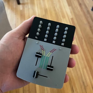 May include: A small, grey, rectangular device with a black panel featuring multiple round buttons. The device has two black knobs with white lines and a colourful graphic of a rainbow with red, yellow, and green dots.