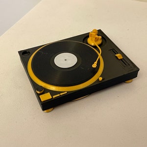 May include: A black and gold record player with a white vinyl record on the turntable. The tonearm is raised and the needle is visible.