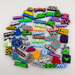 May include: A collection of colorful, small, rubberized shoe charms featuring various music artist logos, band names, and graphic icons in diverse fonts and vibrant colors including neon green, blue, pink, orange, and black on a white background.