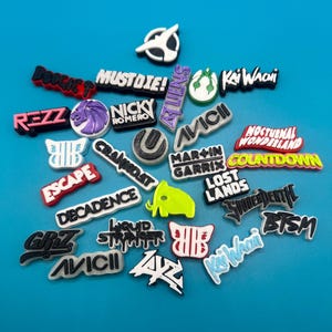 May include: A collection of colorful, printed, plastic charms featuring the names of various music festivals and DJs, including "REZZ", "MUSTDIE!", "KAIWACHI", "AVICII", "MARTIN GARRIX", "COUNTDOWN", "LOST LANDS", "DECADENCE", "CRAMIDAT", "ESCAPE", "NOCTURNAL WONDERLAND", "LIQUID STRANGER", "GRZ", "BTSM", "SHUDDERDEATH", "ZAZ", "NICKY ROMERO", "BOOGIE T".