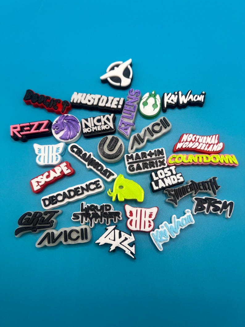 May include: A collection of colorful, printed, plastic charms featuring the names of various music festivals and DJs, including "REZZ", "MUSTDIE!", "KAIWACHI", "AVICII", "MARTIN GARRIX", "COUNTDOWN", "LOST LANDS", "DECADENCE", "CRAMIDAT", "ESCAPE", "NOCTURNAL WONDERLAND", "LIQUID STRANGER", "GRZ", "BTSM", "SHUDDERDEATH", "ZAZ", "NICKY ROMERO", "BOOGIE T".