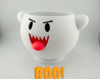 May include: A white, round, 3D-printed planter shaped like a ghost character with two small side arms. It features black oval eyes, a wide red mouth with white teeth, and a protruding red tongue. The orange text BOO! is below the planter.