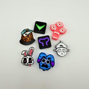 May include: A collection of colorful, cartoonish shoe charms. The charms feature various designs, including a bearded man in a cap, a rabbit, a blue monster, and geometric shapes with symbols. The charms are in shades of pink, blue, black, and white.