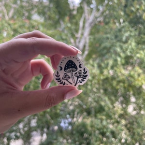 May include: A sticker featuring a black and white mushroom design with crescent moons and leafy accents. The sticker has a whimsical, illustrative style, perfect for decorating laptops, water bottles, or journals.
