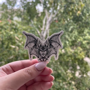 May include: A black and white sticker featuring a detailed illustration of a bat with outstretched wings. The bat has a textured body and detailed wing patterns. The sticker is held against a blurred green background.