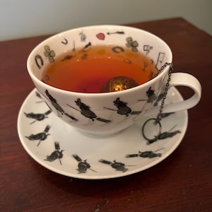 Op de afbeelding: Een witte kop en schotel set met een zwart heksen thema ontwerp. De kop is gevuld met thee en heeft een metalen thee-infuser. De schotel en kop hebben een herhalend patroon van heksen op bezems. De kop heeft ook kleine decoratieve iconen.