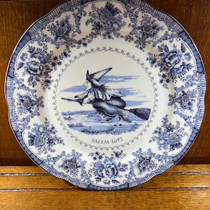 Puede incluir: Un plato de cerámica blanco con un patrón floral azul y blanco. El centro del plato presenta una ilustración en blanco y negro de una bruja volando en una escoba. El texto "SALEM-1692" está impreso debajo de la ilustración.