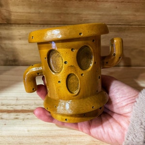 May include: A yellow ceramic cactus planter with a face and small holes. The planter has two arms and a rounded top.