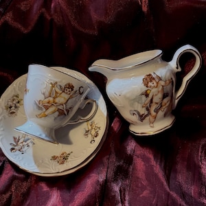 May include: A white porcelain teacup and saucer set with a matching creamer. The set features cherub illustrations and gold trim. Floral designs adorn the saucer. The teacup is tilted, and the creamer has a curved handle.