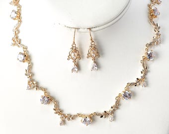 May include: A gold-toned jewelry set featuring a necklace and matching earrings. The necklace and earrings are adorned with clear, sparkling gemstones and delicate leaf-like accents. The necklace is displayed on a white bust.