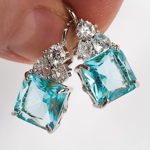 Pode incluir: Brincos de prata pendurados com uma pedra preciosa azul quadrada e detalhes de cristal transparente.