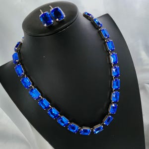 Może przedstawiać: Naszyjnik i pasujące kolczyki z niebieskich kryształów. Naszyjnik wykonany jest ze srebrnego łańcuszka z prostokątnymi niebieskimi kryształami. Kolczyki również są srebrne z prostokątnymi niebieskimi kryształami.