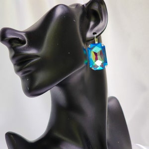 Puede incluir: Un par de pendientes colgantes con una gran piedra rectangular azul iridiscente. Los pendientes son plateados y tienen un diseño sencillo.