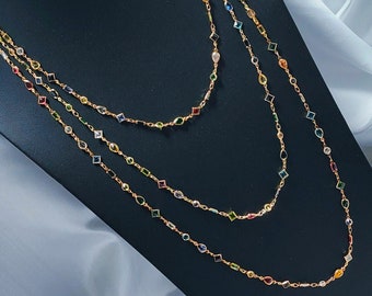 May include: Three gold necklaces with colorful gemstone accents. Each necklace features a delicate chain adorned with various shapes and colors of gemstones, including emerald green, sapphire blue, ruby red, and citrine yellow. The necklaces are displayed on a black surface.
