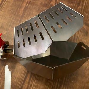 Könnte beinhalten: Ein Bratrostkorb aus Edelstahl mit einem Klappdeckel mit geschlitzten Öffnungen. Der Korb ist mit einem roten Ventil und einer Metallklemme an einem Rohr befestigt. Ein kleines Metallwerkzeug ist ebenfalls vorhanden.