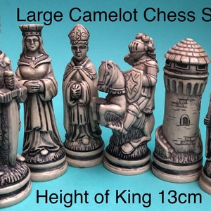 Può includere: Un set di sei pezzi degli scacchi di un grande set di scacchi Camelot. I pezzi sono realizzati in un materiale grigio chiaro e sono dettagliati con intricate sculture. I pezzi includono un re, una regina, un alfiere, un cavallo, una torre e un pedone. Il re è alto 13 cm.  "Large Camelot Chess Set" e "Height of King 13cm" sono stampati sotto i pezzi.