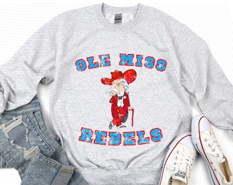May include: Pale grey sweatshirt featuring the text "OLE MISS REBELS" in distressed red and blue lettering. The graphic shows a cartoon of an elderly man in a red suit and hat, holding a cane. White trainers and blue jeans are next to the sweatshirt.