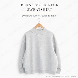 May include: A pale grey mock neck sweatshirt suspended from a wooden hanger. The sweatshirt features a high neckline and long sleeves. Text on the image reads "BLANK MOCK NECK SWEATSHIRT" and "Premium Basic - Ready to Ship". The sweatshirt is unisex fit.