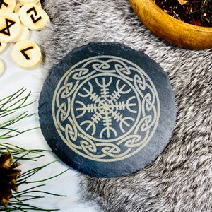 May include: A black slate coaster featuring a gold-coloured Vegvisir symbol encircled by a Celtic knot design.