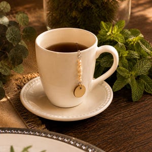 Puede incluir: Una taza de té y un platillo de cerámica blanca que contienen té oscuro. Una cadena de metal plateado cuelga sobre el borde, con una serie de pequeñas cuentas cilíndricas claras y un dije de madera redondo, plano y claro al final.