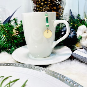May include: A white ceramic mug with a green and white beaded charm hanging from the handle. The charm has a round wooden tag at the bottom.