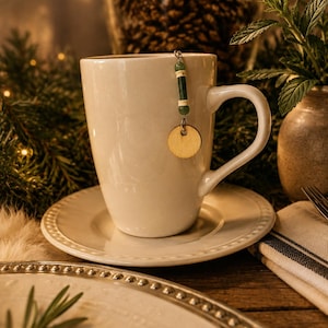 May include: A white ceramic mug sits on a matching saucer. A decorative charm hangs from the rim, featuring a small green cylindrical bead flanked by two smaller green spheres and a circular wooden disc suspended by a silver metal chain.
