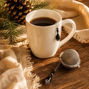 May include: A white ceramic mug filled with dark liquid, featuring a silver chain attachment with a black teardrop bead. Beside it lies a spherical metal tea infuser with a matching silver chain and black teardrop bead charm attached to the end.