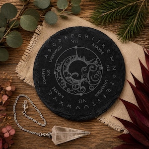 May include: A round black slate board featuring an engraved alphabet, the words YES, NO, MAYBE, UNKNOWN, and GOODBYE, and a central sun and moon design. A clear, faceted crystal pendulum on a silver-toned metal chain is positioned next to the board.