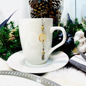 May include: A white ceramic mug with a wooden bead and charm tea infuser hanging from the rim. The mug is sitting on a white saucer.
