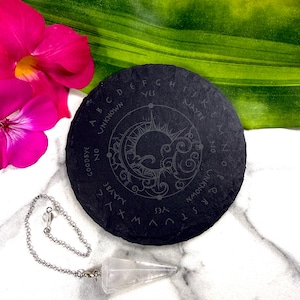 May include: A black slate planchette with engraved letters and symbols, including a sun and moon design. A clear crystal pendulum hangs below. The planchette is surrounded by pink flowers and green leaves.