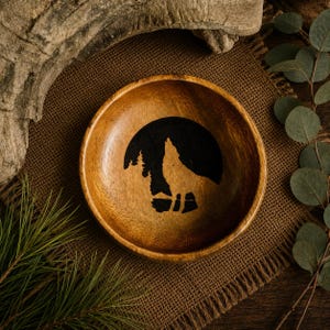 Puede incluir: Un cuenco redondo de madera con un gráfico negro oscuro de un lobo aullando a la luna llena con pinos de fondo. El cuenco tiene un acabado de veta de madera natural de color marrón claro y una superficie interior circular y lisa.