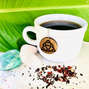 May include: A white ceramic teacup with a black tea infuser shaped like a Celtic knot. The infuser is hanging from a silver chain. The teacup is filled with dark tea and is sitting on a white surface with loose tea leaves scattered around it.