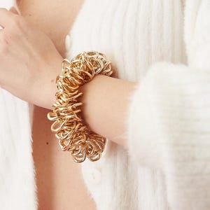 Puede incluir: Un brazalete dorado hecho de anillos de metal entrelazados. El brazalete es grueso y robusto, y se lleva en la muñeca.
