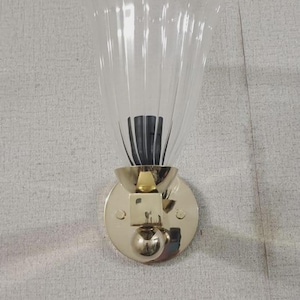 May include: A wall-mounted light fixture with a clear, fluted glass shade and a gold-colored metal base. The shade has a scalloped top edge. The fixture is mounted on a textured, light gray wall.