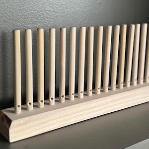 May include: A wooden yarn swift with a rectangular base and numerous vertical dowels. The natural wood color of the swift contrasts with the dark background. This tool is used for winding yarn into a skein.