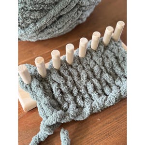 Op de afbeelding: Een close-up van een breiproject in uitvoering. Het project toont een lichtgroen, dik garen dat wordt bewerkt op een houten breiraam. Een bol van hetzelfde garen staat op de achtergrond. Het weefgetouw heeft verschillende houten pinnen.