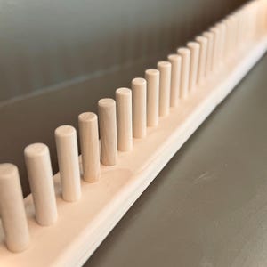 Op de afbeelding: Een houten penweefgetouw met een reeks gelijkmatig verdeelde, cilindrische pennen. De pennen zijn licht van kleur en staan op een rechthoekige basis. Het weefgetouw is gemaakt van onbewerkt hout, dat de natuurlijke nerfstructuur laat zien. De pennen zijn ongeveer 2.5 cm hoog.