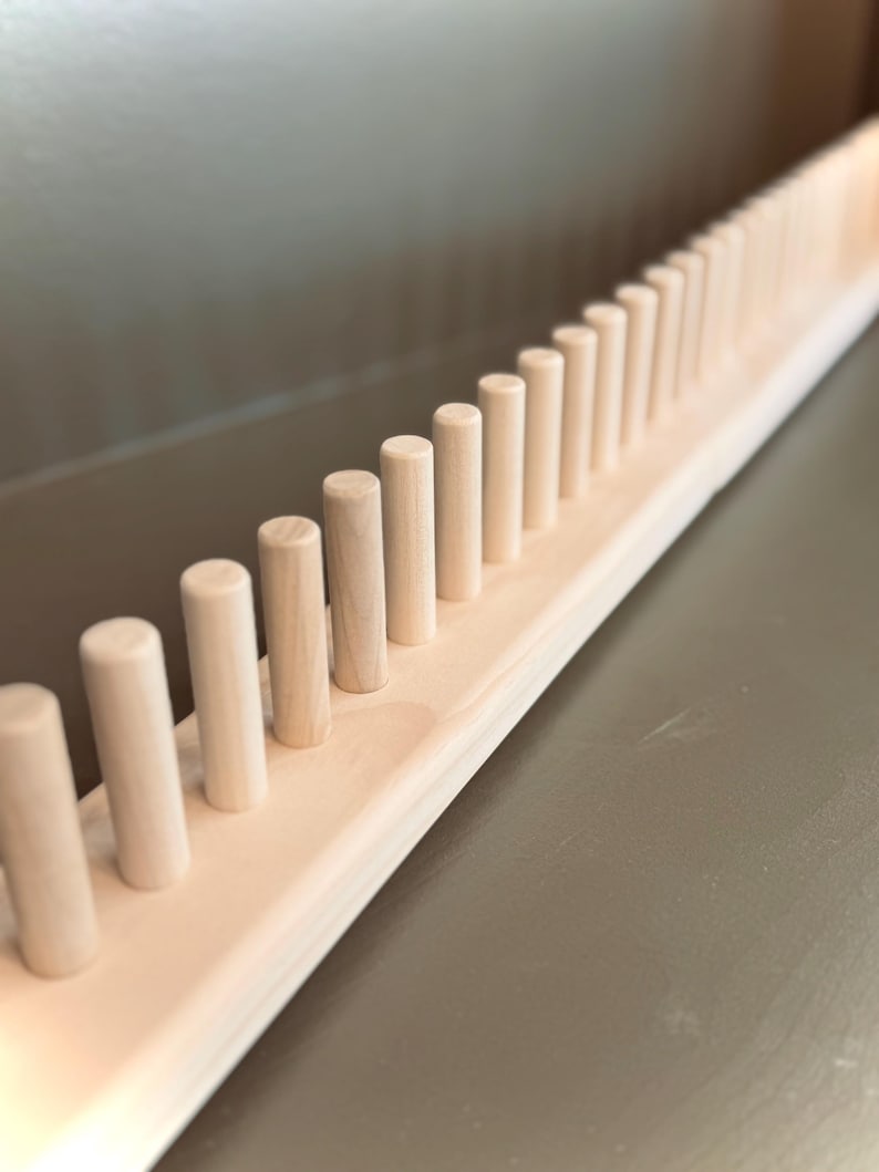 Op de afbeelding: Een houten penweefgetouw met een reeks gelijkmatig verdeelde, cilindrische pennen. De pennen zijn licht van kleur en staan op een rechthoekige basis. Het weefgetouw is gemaakt van onbewerkt hout, dat de natuurlijke nerfstructuur laat zien. De pennen zijn ongeveer 2.5 cm hoog.