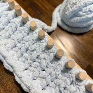 Puede incluir: Una manta de hilo azul claro en proceso sobre un telar de madera. El telar tiene clavijas de madera espaciadas uniformemente. Una madeja de hilo azul claro a juego está cerca. El fondo es una superficie de madera oscura.