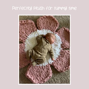 May include: A baby lies on a large, flower-shaped, plush blanket. The blanket has a white center and pink petals. The baby is wearing a brown onesie. The text "Perfectly plush for tummy time" is at the top of the image.