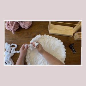May include: A close-up shot of a white, fluffy, round blanket being crafted. A pair of hands is using scissors to cut the yarn. Two balls of pink yarn are in the background, along with a wooden shelf and a staple gun.