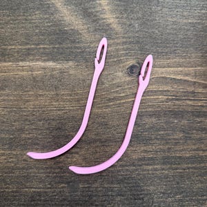 May include: Two pink yarn needles are displayed against a dark wood grain background. The needles have a curved design with an eye at the top for threading yarn. The needles are made of a flexible material.