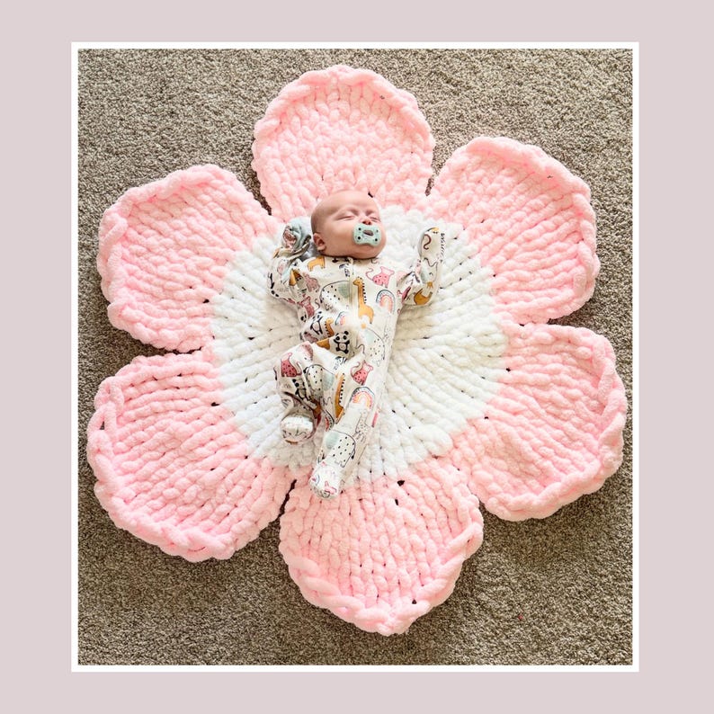 May include: A newborn baby lies on a large, handmade, flower-shaped blanket. The blanket is made of soft, chunky yarn in white and pink. The baby wears a patterned onesie and has a pacifier in their mouth.